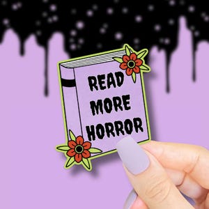 K&ouml;nnte beinhalten: Ein Aufkleber mit einem lila Buch mit den Worten "READ MORE HORROR" in Wei&szlig;. Das Buch ist mit roten und orangefarbenen Blumen verziert. Der Hintergrund ist helllila mit schwarzen Tropfen.