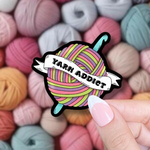 Pode incluir: Um autocolante colorido com uma bola de l&atilde; e uma agulha de croch&ecirc;. A bola de l&atilde; &eacute; listrada com rosa, roxo, verde e amarelo. O texto "YARN ADDICT" est&aacute; em uma faixa. O fundo &eacute; preenchido com bolas de l&atilde;.