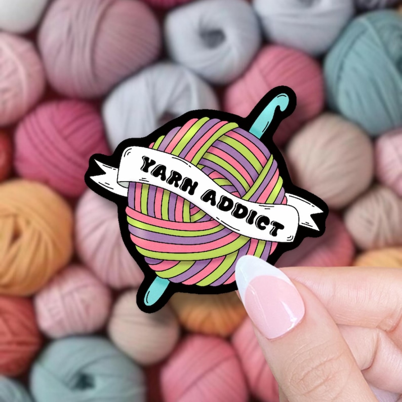 Pode incluir: Um autocolante colorido com uma bola de l&atilde; e uma agulha de croch&ecirc;. A bola de l&atilde; &eacute; listrada com rosa, roxo, verde e amarelo. O texto "YARN ADDICT" est&aacute; em uma faixa. O fundo &eacute; preenchido com bolas de l&atilde;.