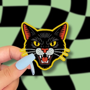 May include: A black cat sticker with a fierce expression, yellow eyes, and a red mouth. The sticker has a yellow border and is held against a green and black checkered background.