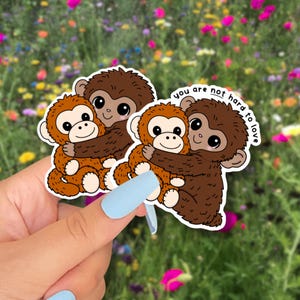 Puede incluir: Dos pegatinas con monos de dibujos animados abrazándose. Un mono es marrón, el otro es naranja. Las pegatinas tienen el texto "you are not hard to love". El fondo es un campo de flores coloridas.