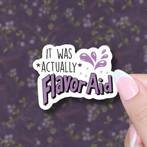 May include: A white sticker with the text "IT WAS *ACTUALLY* Flavor Aid" in black and purple. The sticker has a purple splash design and is held by a hand with a French manicure. The background is a floral pattern.