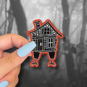 May include: A whimsical sticker featuring a black and white illustration of a house with chicken legs, a chimney, and stars. The house has an orange outline and is set against a blurred forest background.