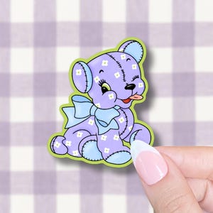 May include: A purple teddy bear sticker with a light blue bow and accents. The bear has white flower details and is winking with its tongue sticking out. The sticker has a green border and is on a checkered background.