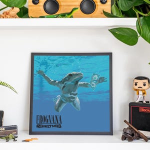 May include: A square framed art print featuring a blue underwater scene with a frog swimming towards a dollar bill. The text FROGVANA and RIBBITMIND is printed in black at the bottom of the image. The print is enclosed in a simple black frame.