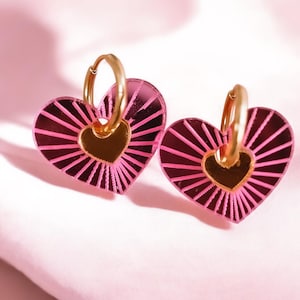 Può includere: Un paio di orecchini a forma di cuore. Gli orecchini presentano cerchi dorati e ciondoli a forma di cuore rosa con cuori dorati al centro. Il design del cuore ha linee radiali.