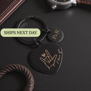 May include: Black heart-shaped keychain with a hand sign and the words "I love you" in a cursive font. A small round tag with the initials "P&R" is attached. The keychain is on a black surface, with a watch and leather accessories in the background.