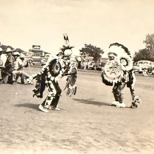 May include: Black and white vintage photograph of a Native American Shield Dance at a Kickapoo Pow-Wow in Horton, Kansas. Two dancers in traditional regalia perform in front of a crowd and vintage cars. The text "Shield Dance Kickapoo Pow-Wow Horton Kans." is at the bottom.