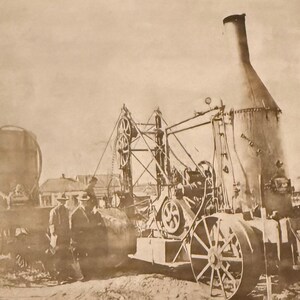 May include: Sepia-toned photograph of an antique steam engine with large wheels, a tall smokestack, and intricate machinery. Several people stand near the engine, and a building is visible in the background. The image is framed with a decorative border.