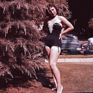 May include: A vintage black swimsuit with white trim and a decorative design on the bodice. The one-piece swimsuit is worn with white heels. The image is taken outdoors with a green lawn and cars in the background.