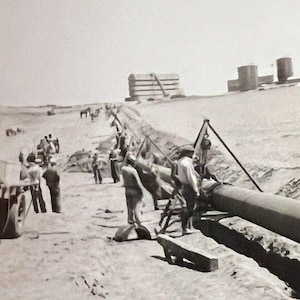 Könnte beinhalten: Schwarz-Weiß-Fotografie einer Baustelle. Arbeiter installieren ein großes Rohr in einem Graben. Gebäude und Ausrüstung sind im Hintergrund zu sehen. Das Bild ist eine Vintage-Fotografie.