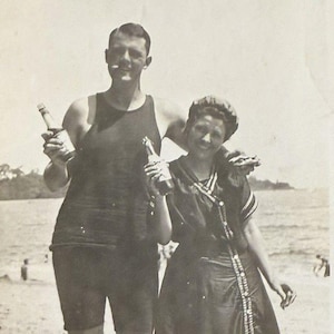 May include: Vintage black and white photograph of a man and a woman on a beach. The man wears a dark swimsuit and holds a bottle. The woman wears a dark dress with a decorative trim and holds a bottle. The background shows the ocean and sky.