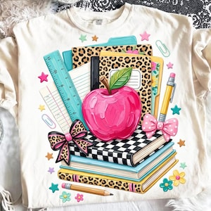 May include: A cream-coloured t-shirt featuring a graphic print of school supplies including a bright pink apple, a stack of books with leopard print and checkered covers, a teal ruler, a yellow pencil, and pink bows, surrounded by small colourful stars.