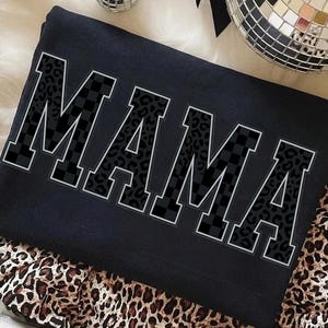 Può includere: Una felpa nera con la scritta "MAMA" in lettere grandi e in grassetto. Le lettere presentano un design unico, con una combinazione di stampa leopardata e motivi a scacchi. La felpa è piegata e appoggiata su una superficie con stampa leopardata.