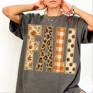 May include: A charcoal gray short-sleeve t-shirt featuring the word FALL in large block letters. Each letter has a distinct pattern: the F has a floral print, the A has a leopard print, the L has an orange gingham check, and the L has polka dots.