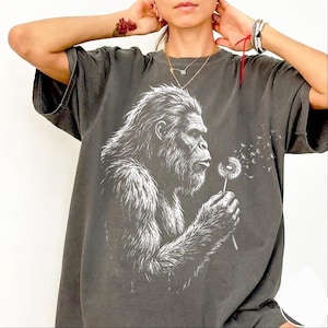May include: Dark gray t-shirt featuring a white graphic of a Bigfoot blowing on a dandelion. The Bigfoot is detailed with fur and facial features. The dandelion's seeds are blowing away. The shirt has short sleeves and a relaxed fit.