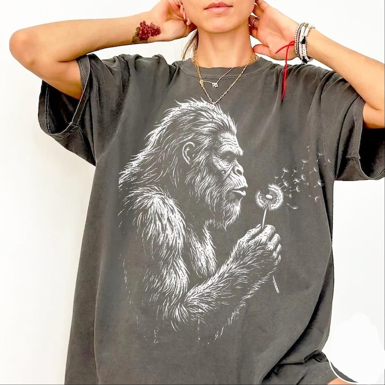 May include: Dark gray t-shirt featuring a white graphic of a Bigfoot blowing on a dandelion. The Bigfoot is detailed with fur and facial features. The dandelion's seeds are blowing away. The shirt has short sleeves and a relaxed fit.