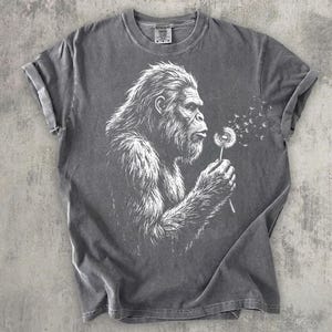 May include: Gray t-shirt featuring a white illustration of a Bigfoot blowing on a dandelion. The Bigfoot is depicted with detailed fur and facial features. The dandelion seeds are blowing away from the Bigfoot. The shirt has rolled-up sleeves.