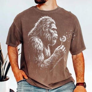 May include: Brown t-shirt featuring a white illustration of a Bigfoot blowing on a dandelion. The graphic design shows the Bigfoot's profile, with detailed fur and facial features. The t-shirt has short sleeves and a relaxed fit.
