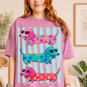 Puede incluir: Una camiseta malva con un estampado gráfico de tres perros salchicha con gafas de sol. El perro de arriba es rosa con lunares amarillos, el del medio es azul con lunares amarillos y el de abajo es rosa con lunares amarillos.