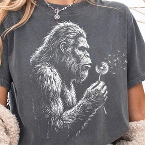May include: Dark gray t-shirt featuring a white graphic of a Bigfoot blowing dandelion seeds. The Bigfoot is depicted in a detailed, artistic style. The shirt has short sleeves and a relaxed fit, suitable for casual wear.