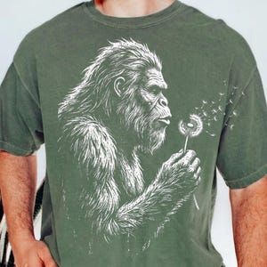 May include: Olive green t-shirt featuring a white illustration of a Bigfoot blowing on a dandelion. The Bigfoot is depicted in profile, with detailed fur and facial features. The dandelion seeds are blowing away from the flower.