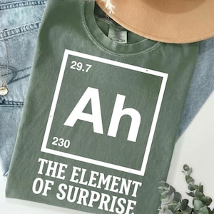 Puede incluir: Una camiseta de manga corta color verde oliva con un gráfico blanco diseñado como una casilla de elemento de la tabla periódica. El gráfico muestra las letras Ah, los números 29.7 y 230, y el texto THE ELEMENT OF SURPRISE en letras mayúsculas.