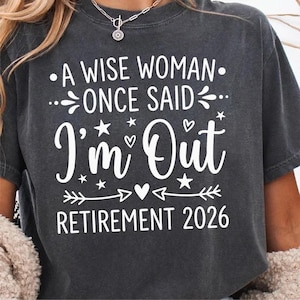 May include: Dark gray t-shirt with white text: "A wise woman once said, I'm out, Retirement 2026." The design includes stars, hearts, and arrows. The shirt is a casual wear item.