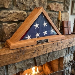 May include: A wooden triangular flag display case with a glass front, holding a folded American flag. The flag has white stars on a blue field and red and white stripes. A small plaque reads "SGT JOHN A. SMITH". The case sits on a wooden mantel above a fireplace.