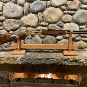 May include: A lever-action rifle with a walnut stock and forend, brass receiver, and blued barrel. The rifle is displayed on a wooden stand in front of a stone wall. The firearm is a classic hunting rifle.