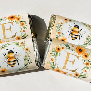 May include: Four wrapped candy bars with bee-themed labels. The labels feature a watercolor design of bees, sunflowers, and the letter "E". The wrappers are silver.