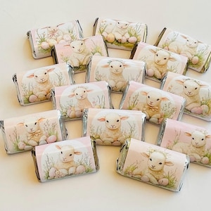 May include: A collection of individually wrapped chocolate bars. Each bar features a pastel pink and white design with a lamb illustration, Easter eggs, and floral accents. The wrappers are silver and gold.