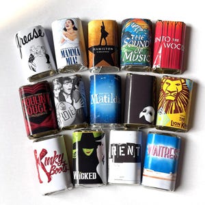 May include: A collection of theater-themed matchbox covers. The covers feature titles like "Grease," "Mamma Mia," "Hamilton," "The Sound of Music," "Into the Woods," "Moulin Rouge," "Bye Bye Birdie," "Matilda," "The Phantom of the Opera," "The Lion King," "Kinky Boots," "Wicked," "Rent," and "Waitress."