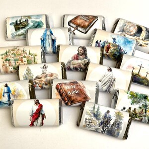 May include: A collection of individually wrapped chocolate bars with watercolor illustrations depicting religious scenes. The images include Jesus, the Holy Bible, and other biblical events. The bars are arranged on a white surface.