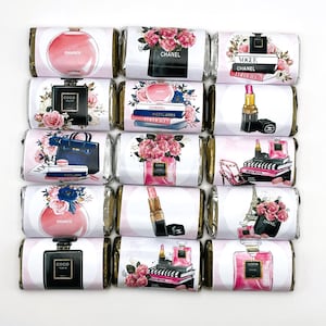 May include: Fifteen rectangular chocolate bars wrapped in white paper with fashion-themed illustrations including perfume bottles, lipsticks, handbags, high heels, stacks of books, and floral arrangements in shades of pink, black, and gold.