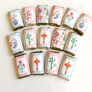 May include: A collection of wrapped chocolate bars with decorative designs. Each bar features a different illustration, including flowers, birds, and traditional Asian motifs. The wrappers have pink and blue borders.