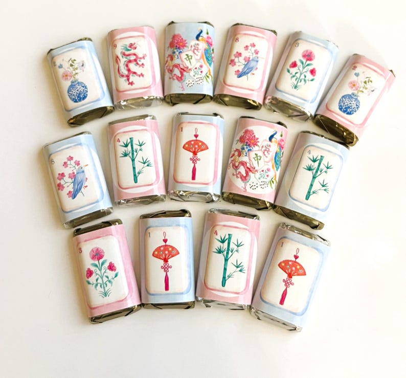Puede incluir: Una colecci&oacute;n de barras de chocolate envueltas con dise&ntilde;os decorativos. Cada barra presenta una ilustraci&oacute;n diferente, incluyendo flores, p&aacute;jaros y motivos asi&aacute;ticos tradicionales. Los envoltorios tienen bordes rosas y azules.