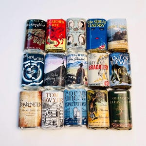 May include: A collection of 15 book-themed beverage can covers, each featuring a classic novel's title and cover art. Titles include "To Kill a Mockingbird," "The Great Gatsby," and "Fahrenheit 451." The cans are arranged on a white surface.