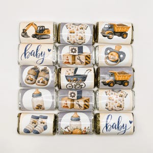 May include: A collection of fifteen rectangular chocolate bars wrapped in foil, each featuring watercolor illustrations of construction vehicles, baby items, and the word baby in blue script, arranged in a grid on a plain white background.