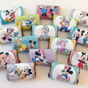 May include: A collection of individually wrapped chocolate bars featuring various Disney characters. The wrappers display images of Mickey Mouse, Minnie Mouse, Donald Duck, and other characters in colorful designs. The bars are arranged on a white surface.
