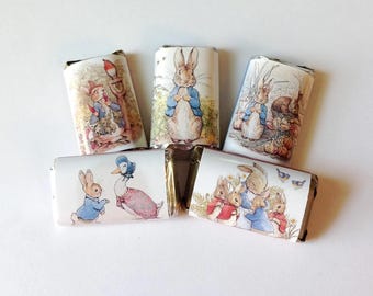 May include: Five chocolate bars with illustrations of Peter Rabbit and other characters from Beatrix Potter's stories. The wrappers feature colorful images of rabbits, ducks, and other animals in a whimsical, storybook style. The bars are arranged on a white surface.