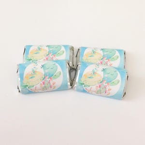 May include: Four individually wrapped candy bars with light blue wrappers. Each wrapper features a watercolor illustration of two turtles, one yellow and one teal, with floral accents. The turtles are inside a white circle.