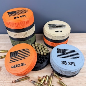 May include: A collection of colorful ammunition storage containers with an American flag design. The containers are orange, tan, green, and blue, with text indicating the caliber: 38 SPL, 9MM, and 40 CAL. Loose bullets are scattered around.