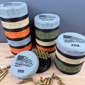 May include: A collection of ammunition storage containers in various colors, stacked and arranged on a wooden surface. Each container is cylindrical with a screw-on lid, labeled with caliber information and the phrase "We The People" alongside an American flag design. Several bullets are scattered around.