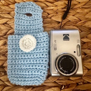 May include: A light blue crocheted camera case with a white flower accent beside a silver Kodak digital camera. The camera has a black lens and the text "4X WIDE" and "FULL HD". The case is on a woven surface.