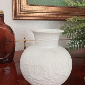 May include: A white matte ceramic vase with a spherical body and a flared rim. The surface features a raised, embossed floral pattern covering the exterior, including various flower shapes and leaves, with a decorative band around the neck.
