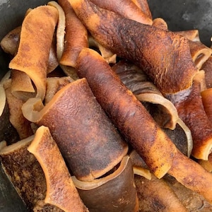 May include: Close-up of a pile of fried pork rinds, with a mix of light tan and dark brown colors. The rinds have a curled shape and a crispy texture, with some pieces appearing translucent.