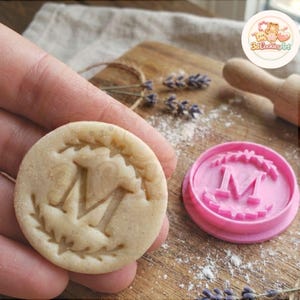 Peut inclure: Un biscuit rond avec la lettre "M" en relief au centre, entouré d'une bordure décorative. À côté, un emporte-pièce rose avec le même motif. Le biscuit est tenu dans une main.