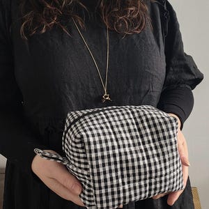 Könnte beinhalten: Eine Kosmetiktasche mit schwarz-weißem Vichy-Muster und Reißverschluss. Die Tasche wird vor einer Person gehalten, die ein schwarzes Kleid und eine goldene Halskette mit einem kleinen Anhänger trägt.