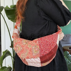 Könnte beinhalten: Eine halbmondförmige Hobo-Tasche mit einem Patchwork-Design aus verschiedenen roten, orangen und cremefarbenen Stoffmustern, darunter Paisley-, Blumen- und geometrische Motive. Die Tasche hat einen breiten, integrierten Schulterriemen und eine weiche, gesteppte Textur.